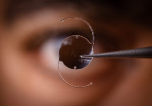 Intraocular lens held by tweezers with a blurred eye in the background.